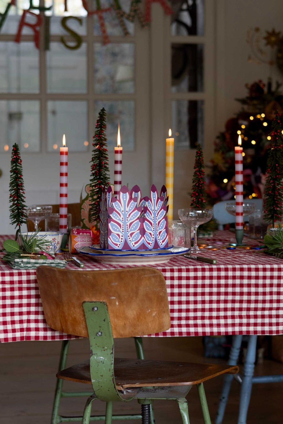 A festive holiday table setting featuring a central red and white patterned cake on a blue-rimmed plate, surrounded by red and white striped taper candles, miniature green trees, and a red-and-white checkered tablecloth.