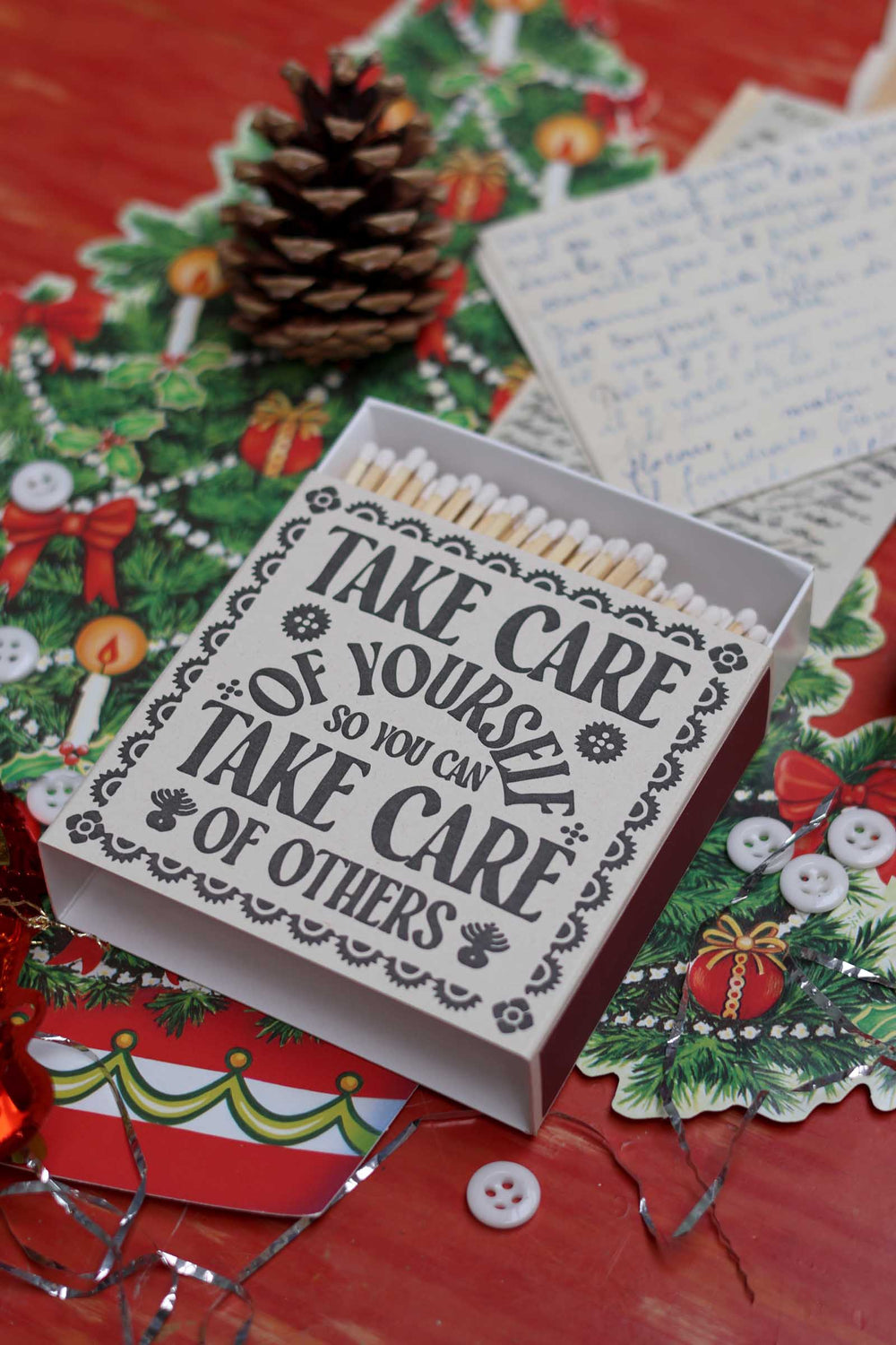 A Christmas-themed matchbox with the quote 'TAKE CARE OF YOURSELF SO YOU CAN TAKE CARE OF OTHERS' printed on the lid, shown open with white matches inside. The matchbox is styled on a red surface with a pinecone, holiday paper, and a handwritten note.