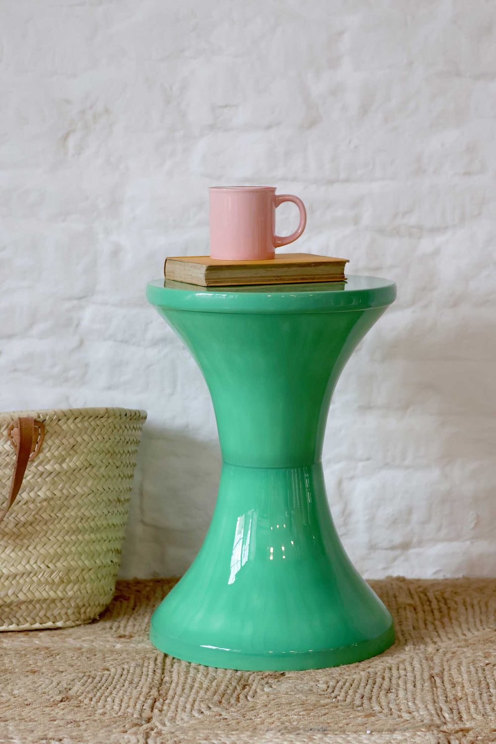 Green side table with a pink mug and book on a textured surface and white wall background