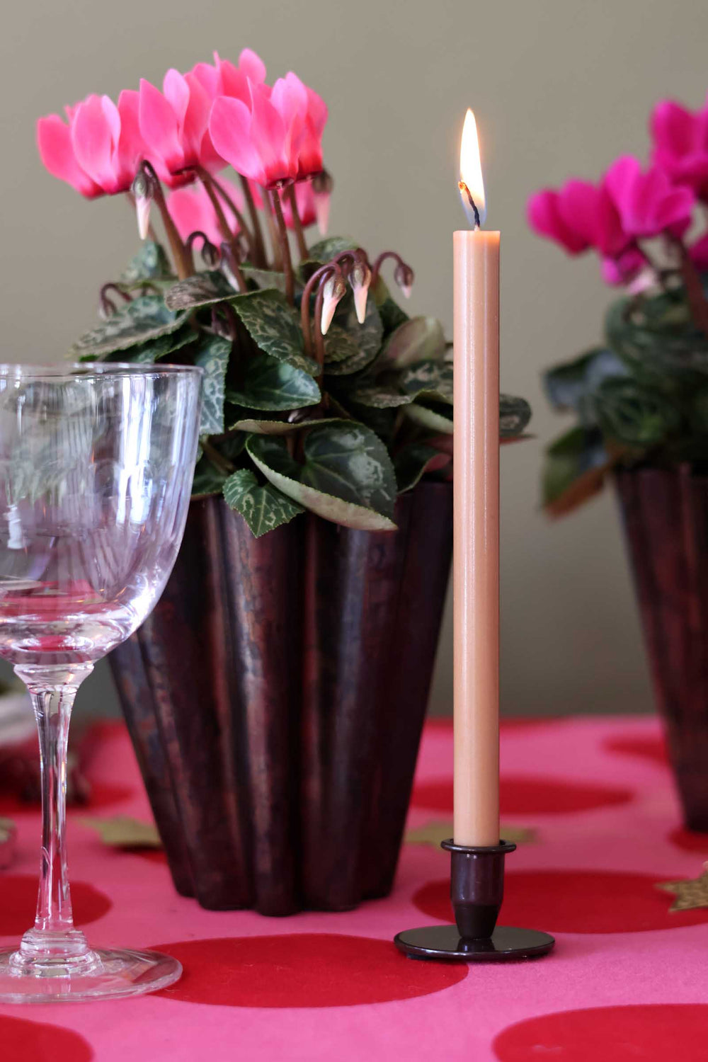 A lit beige taper candle in a black metal holder stands on a pink tablecloth beside a wine glass and potted pink cyclamen flowers.