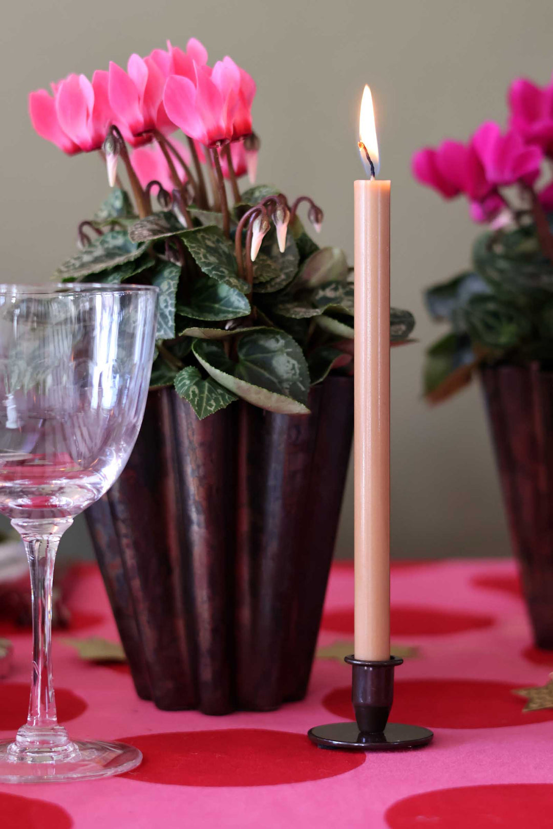 A lit beige taper candle in a black metal holder stands on a pink tablecloth beside a wine glass and potted pink cyclamen flowers.
