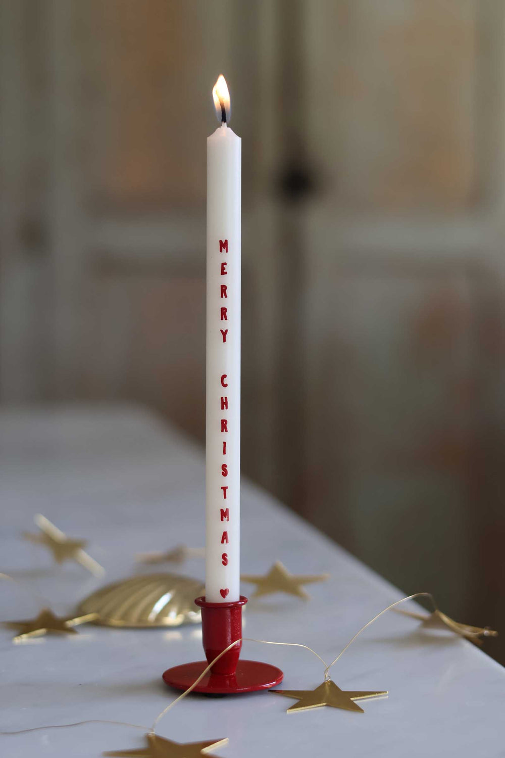 A single white taper candle with 'MERRY CHRISTMAS' printed vertically in red, lit and held in a red holder on a tabletop with gold star decorations in a softly blurred indoor setting.