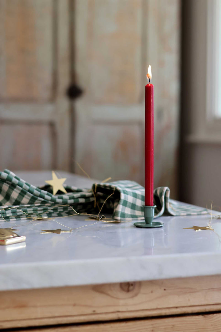 A lit red taper candle in a dark green metal holder sits on a white marble tabletop, with a green-and-white checkered cloth and small gold star decorations in the softly blurred background.