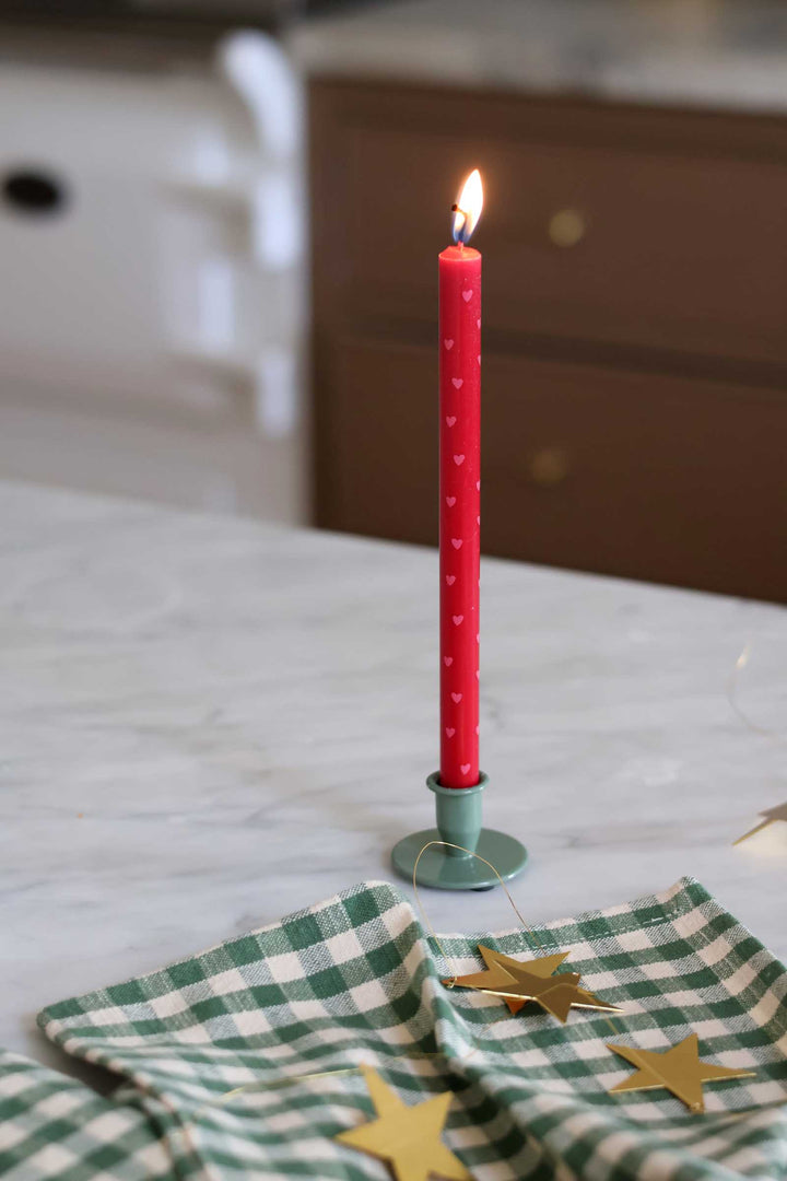 A lit red taper candle with small white heart patterns stands in a mint green metal holder on a marble countertop. A green-and-white checkered cloth with gold star decorations is in the foreground, with a softly blurred kitchen background.