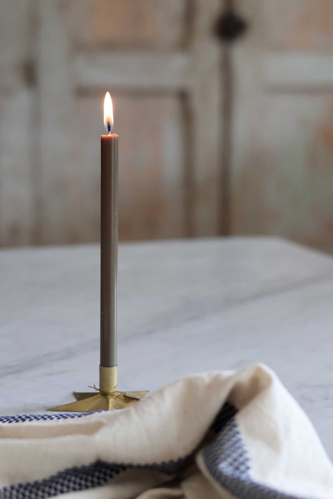 A single lit taper candle in a matte taupe-brown finish with a gold-toned base, set on a light marble tabletop with a cream and navy checkered cloth in the foreground and a softly blurred rustic interior background.