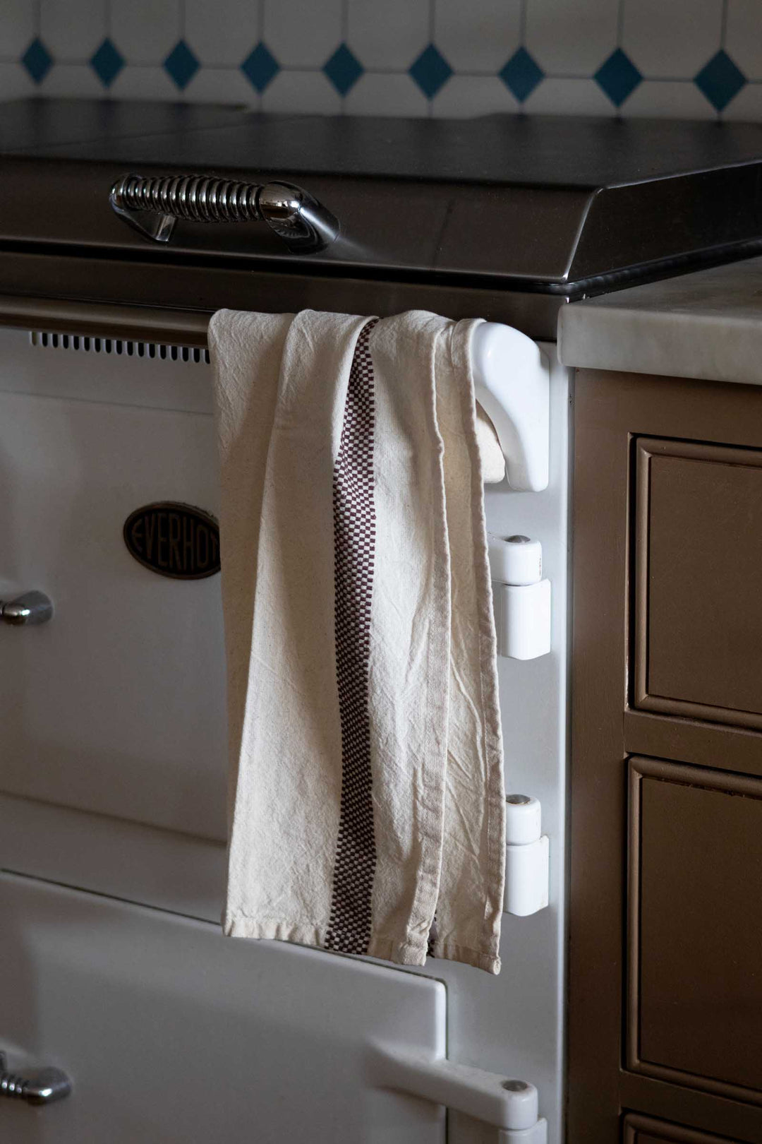 A cream-colored kitchen towel with a dark brown vertical stripe hangs from a white hook on a vintage cream Aga stove in a kitchen setting.