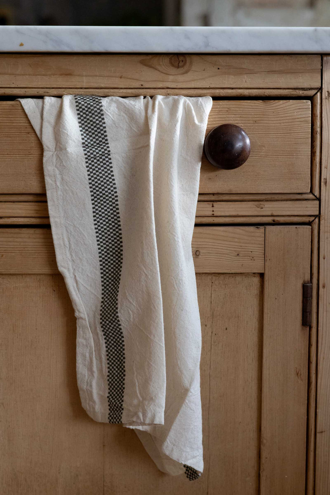 An off-white linen kitchen towel with a vertical black checkered stripe hangs from a dark wooden cabinet knob in a rustic kitchen setting.