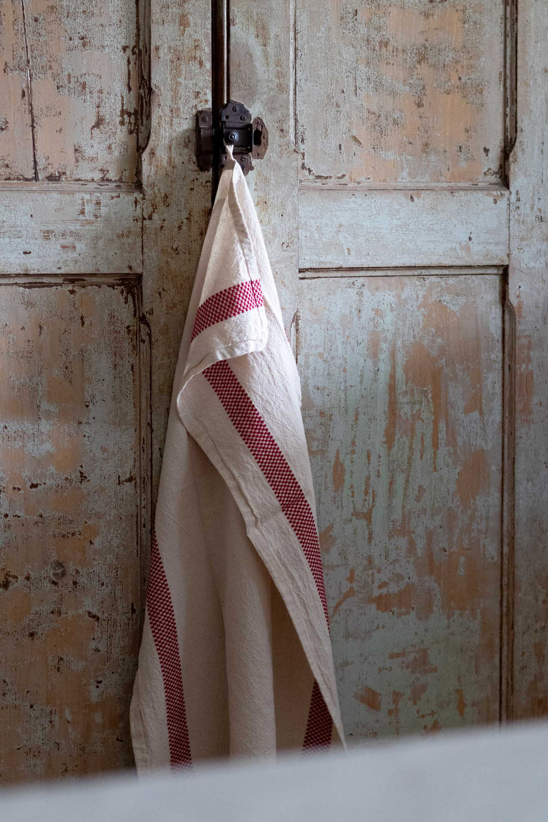 A cream-colored kitchen towel with red stripes hanging from a dark metal hook on a distressed, weathered wooden door.