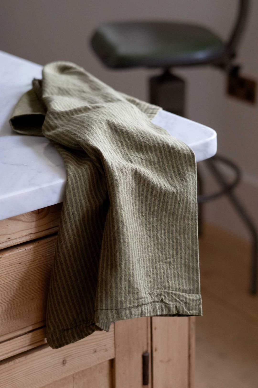 A close-up of olive green striped fabric, likely a towel or garment, draped over the edge of a white countertop in a kitchen setting with wooden cabinets and a chair in the background.