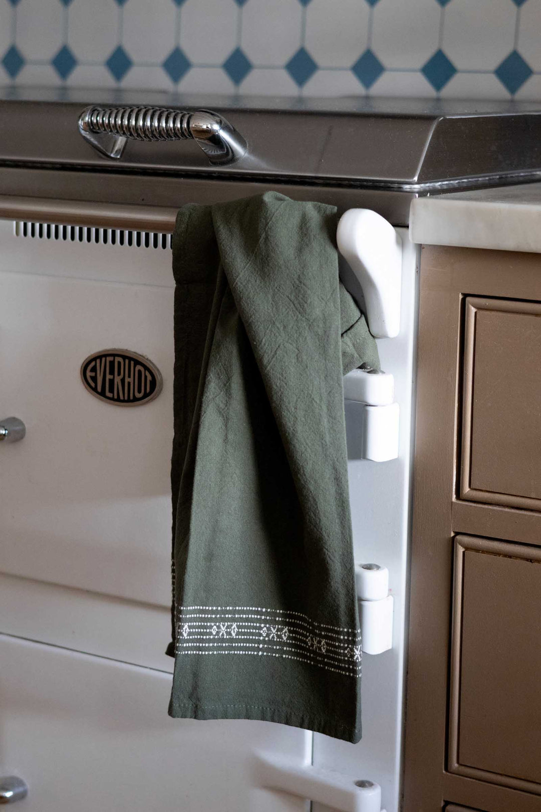 An olive green kitchen towel with white embroidered borders hangs from a white rack attached to a cream-colored Aga stove in a kitchen setting.