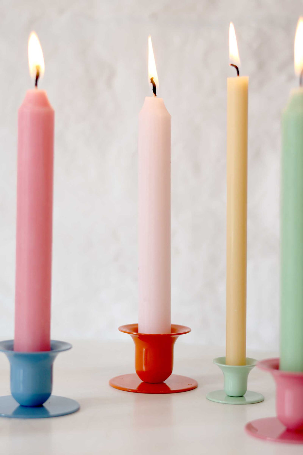 Four lit candles in pastel colors on colorful holders against a light background