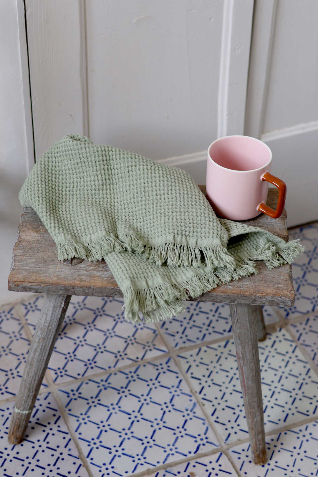 Green woven blanket and pink mug on a wooden stool with a white wall and patterned floor in the background