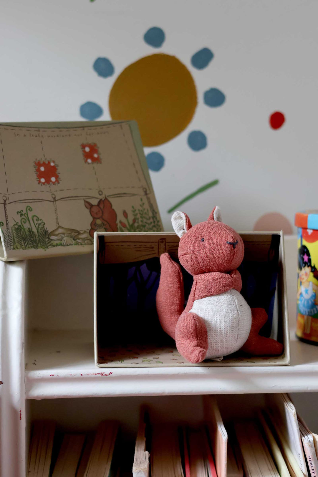 Pink plush toy in a box with children's books and a colorful wall decoration in the background