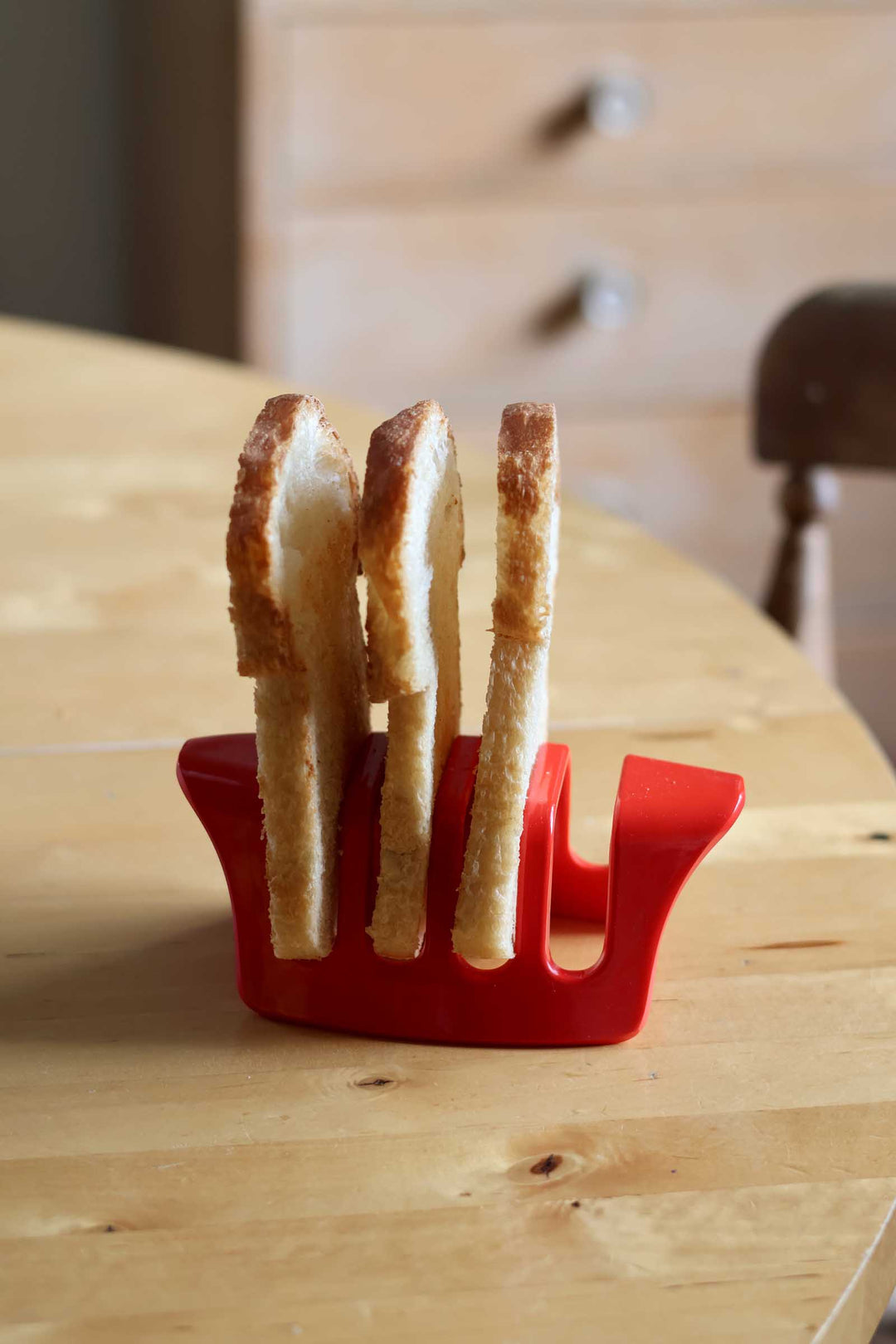 A red plastic toast holder with four slots, holding three slices of toasted bread, set on a light wood table with a blurred background.