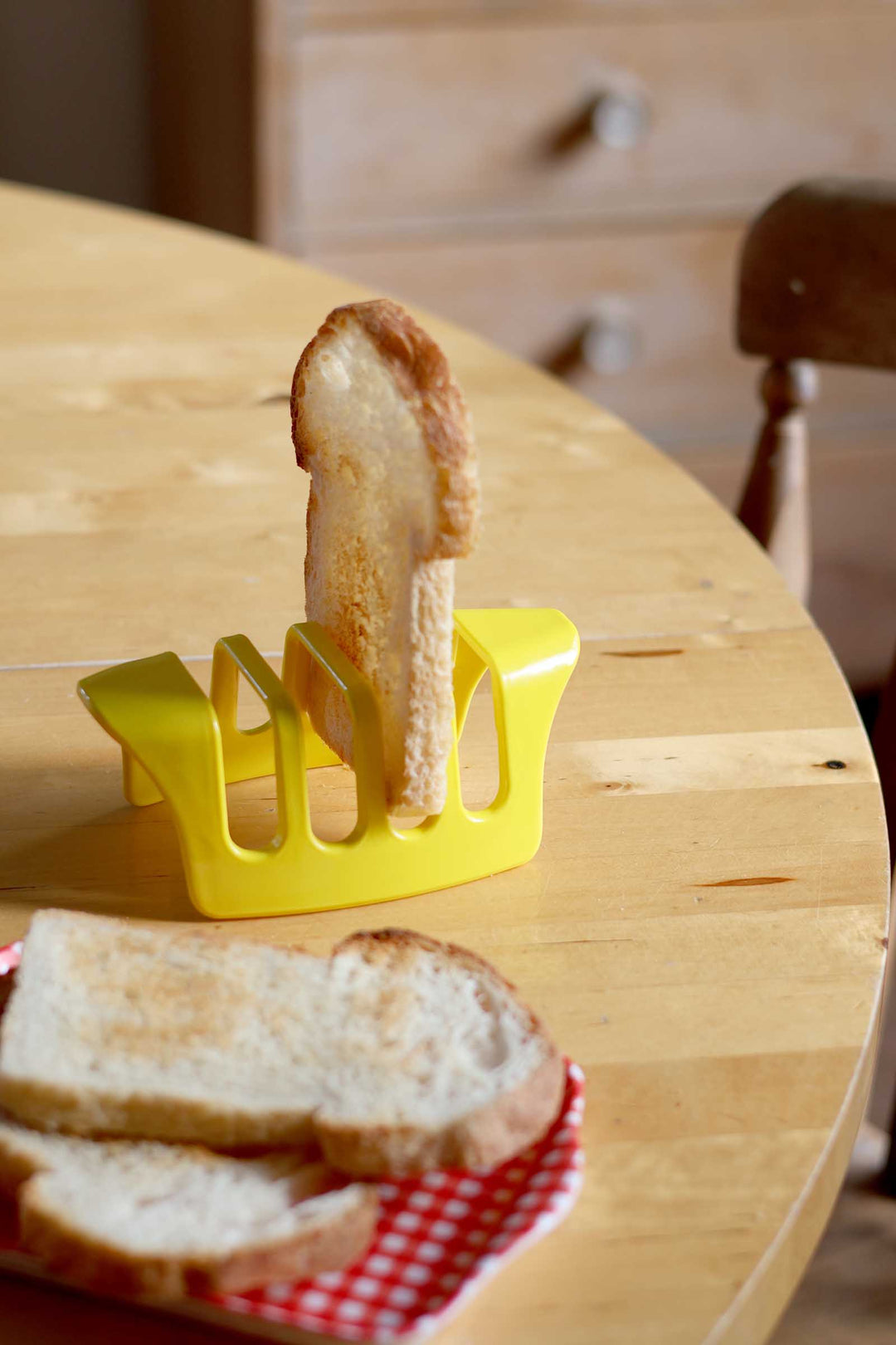 A yellow toast rack holding a slice of toasted bread upright on a wooden table, with additional slices on a red-and-white checkered tray nearby.