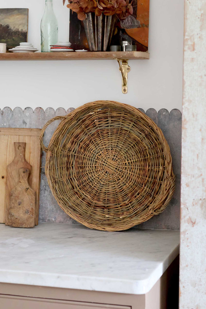 A round woven wicker basket with a handle, displayed on a kitchen counter against a gray scalloped backsplash.