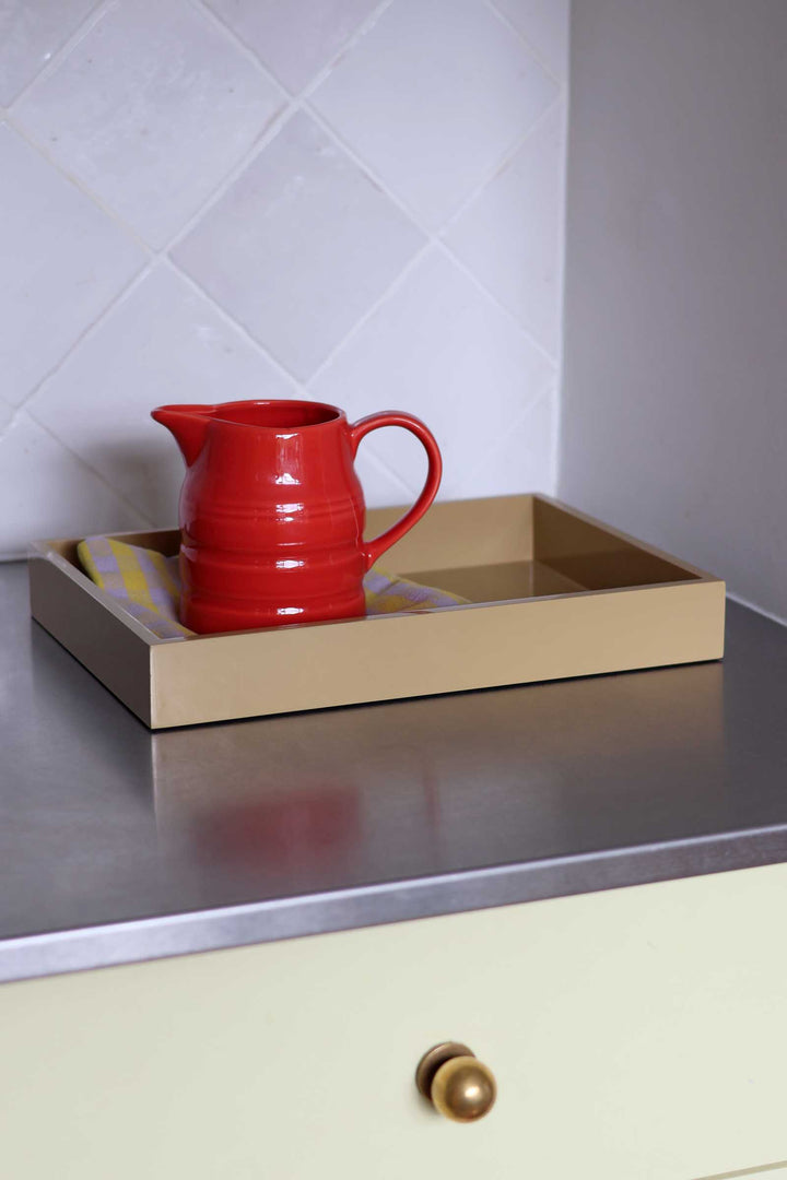 A glossy red ceramic pitcher resting inside a beige rectangular tray on a stainless steel countertop, with a cream-colored cabinet drawer and a white tiled backsplash in the background.