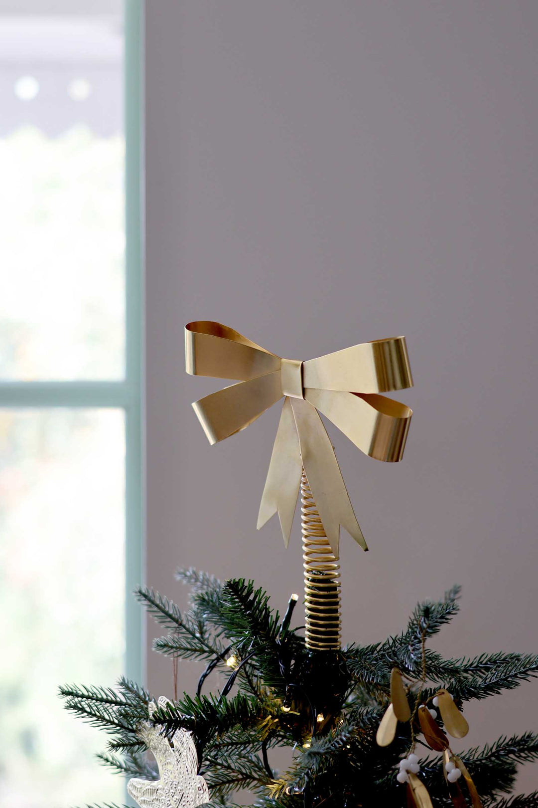 A metallic gold bow tree topper with a spring-style base, mounted on a green Christmas tree branch, set against a soft gray wall and a bright window.