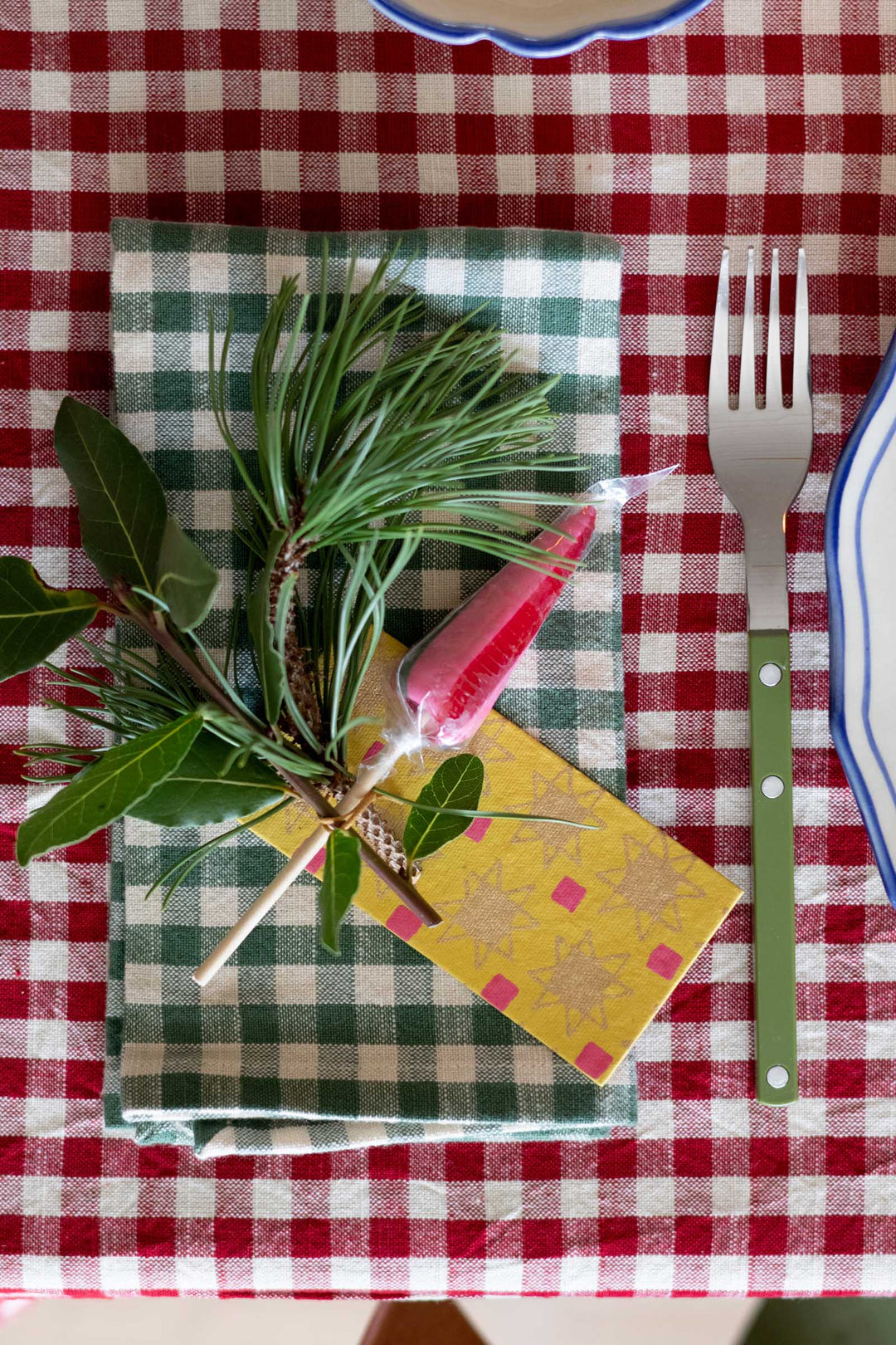 A festive holiday table setting featuring a green and white checkered napkin, a pine sprig with green leaves, a red decorative candy cane ornament, a yellow patterned paper tag, and a green-handled metal fork on a red and white checkered tablecloth.