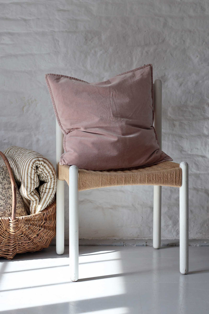 A dusty rose velvet throw pillow with tassel trim sits on a white-framed chair with a woven seat, next to a wicker basket with folded blankets, against a white textured wall.