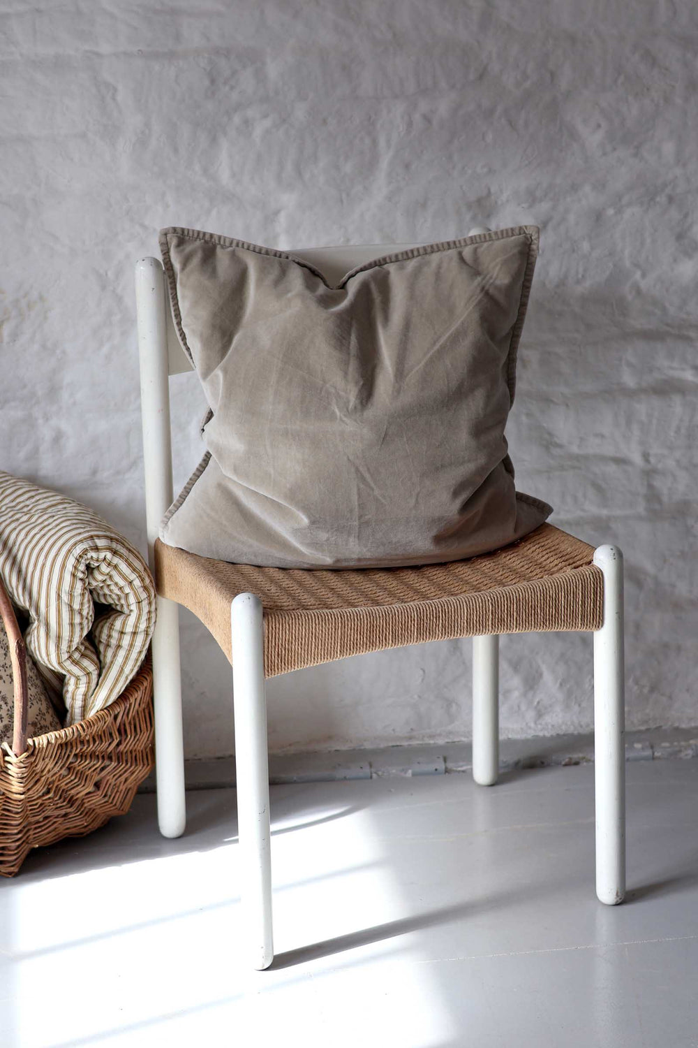 A beige linen throw pillow with a decorative stitched border rests on a white-framed chair with a woven seat, set against a textured gray plaster wall.
