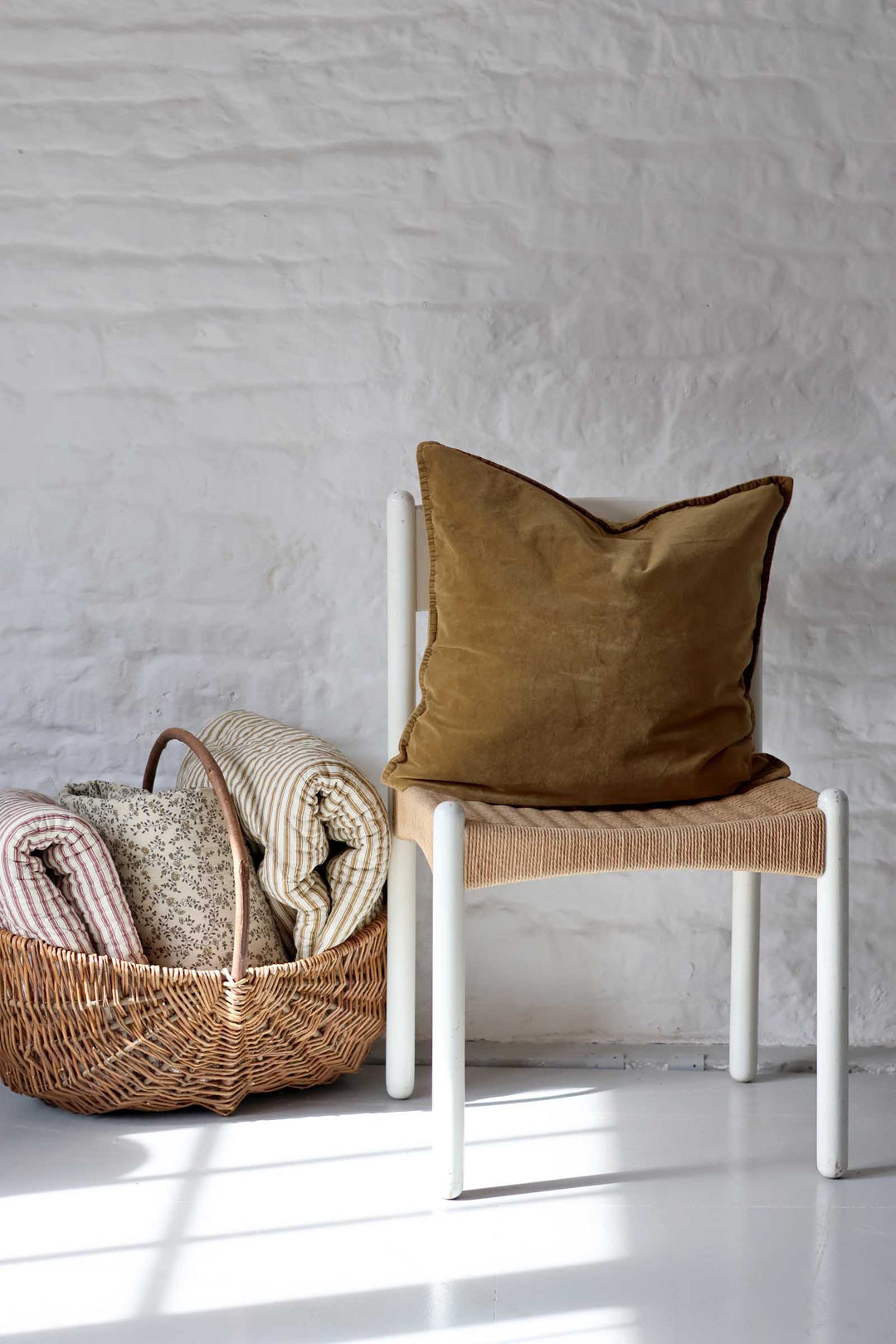 A brown velvet throw pillow with a piped edge sits on a white-framed chair with a woven seat, next to a wicker basket containing folded blankets, set against a white brick wall.