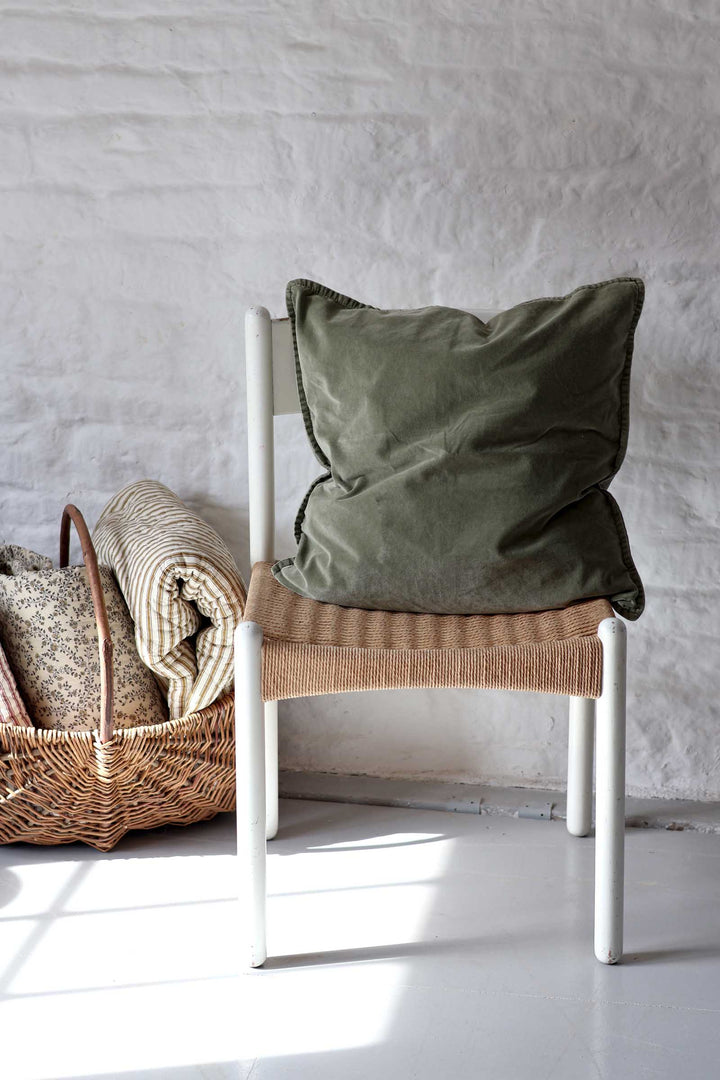 An olive green velvet throw pillow resting on a white-framed woven chair, set against a white brick wall with a wicker basket and folded blankets on the left.