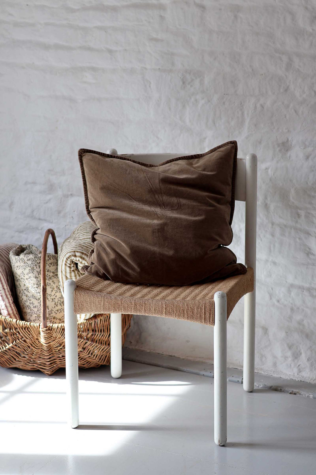 A brown velvet throw pillow resting on a white wooden chair with a woven seat, set against a white textured wall.