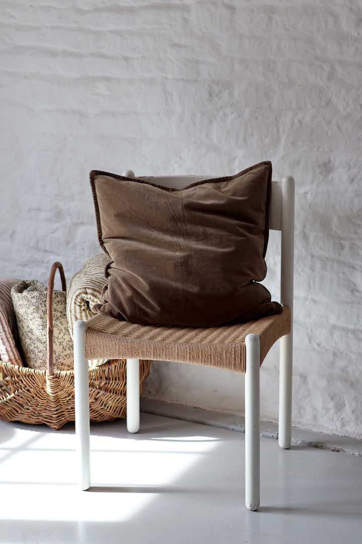 A brown velvet throw pillow resting on a white wooden chair with a woven seat, set against a white textured wall.
