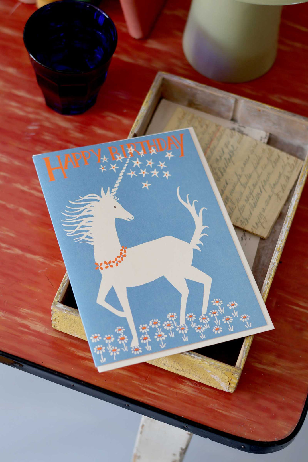 Unicorn birthday card on a wooden surface with a blue glass and white vase in the background.