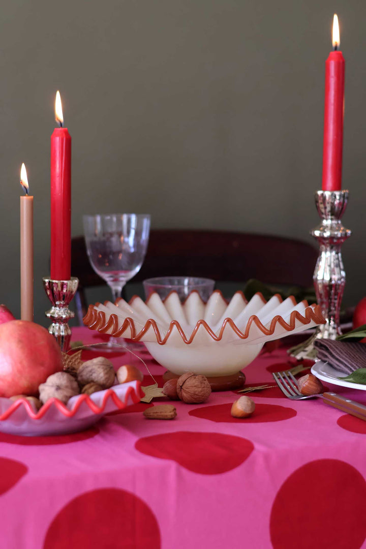 A styled dining table setting featuring a vibrant pink tablecloth with large red polka dots, a white scalloped bowl with a red zigzag trim, red candles in silver candlesticks, walnuts, and a red apple.