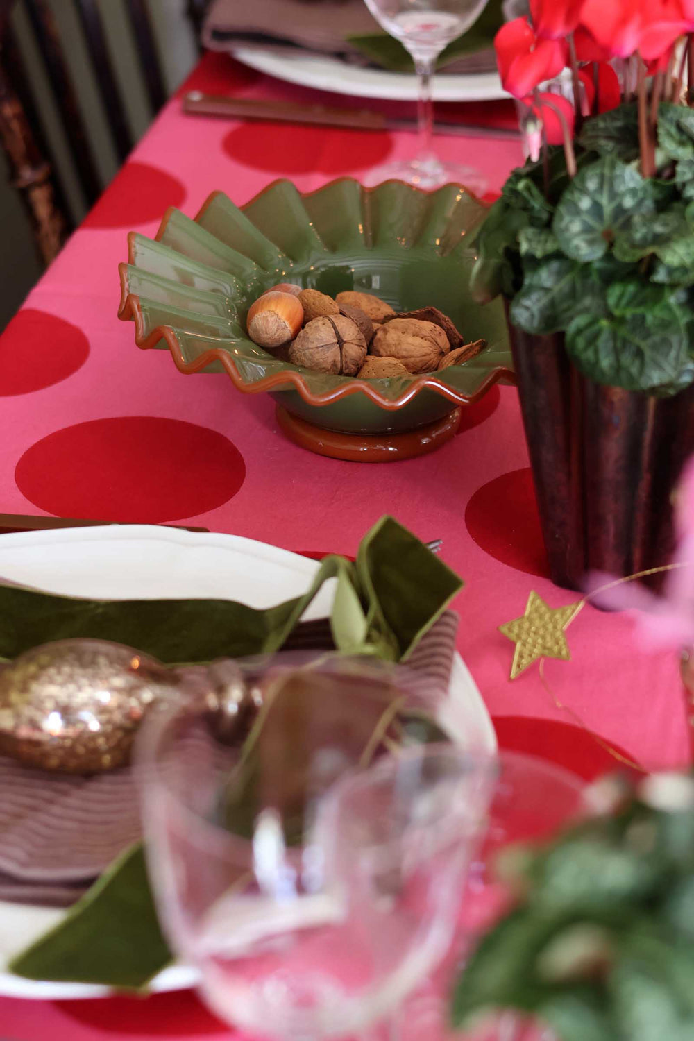 A close-up of a styled dining table setting featuring a vibrant pink tablecloth with large red polka dots. A green scalloped ceramic bowl filled with walnuts sits in the center, surrounded by plates, green leaves, and a potted plant with red flowers.
