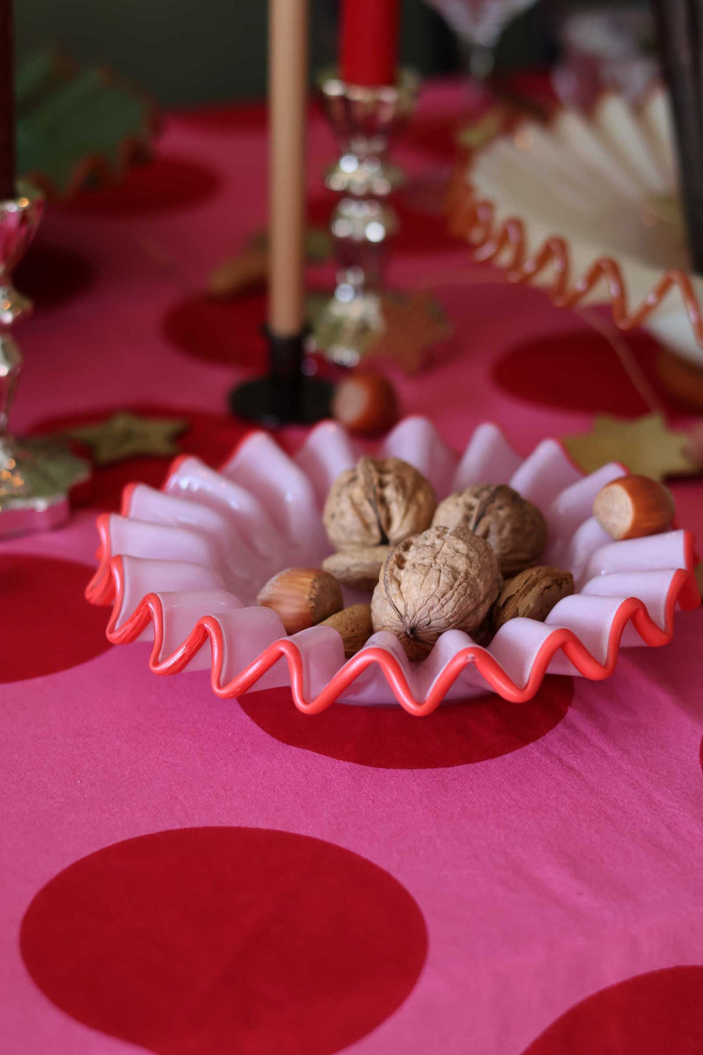 A pink and red wavy-rimmed bowl filled with walnuts and hazelnuts, set on a pink tablecloth with red polka dots and blurred holiday decorations in the background.