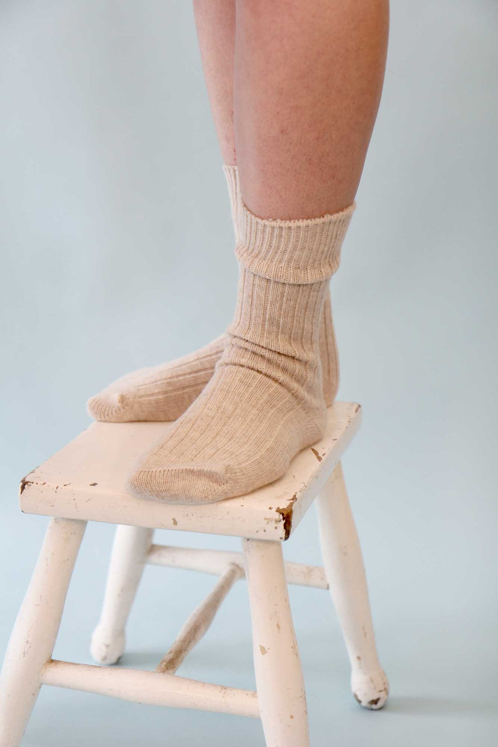 Person wearing beige socks on a small wooden stool against a light gray background