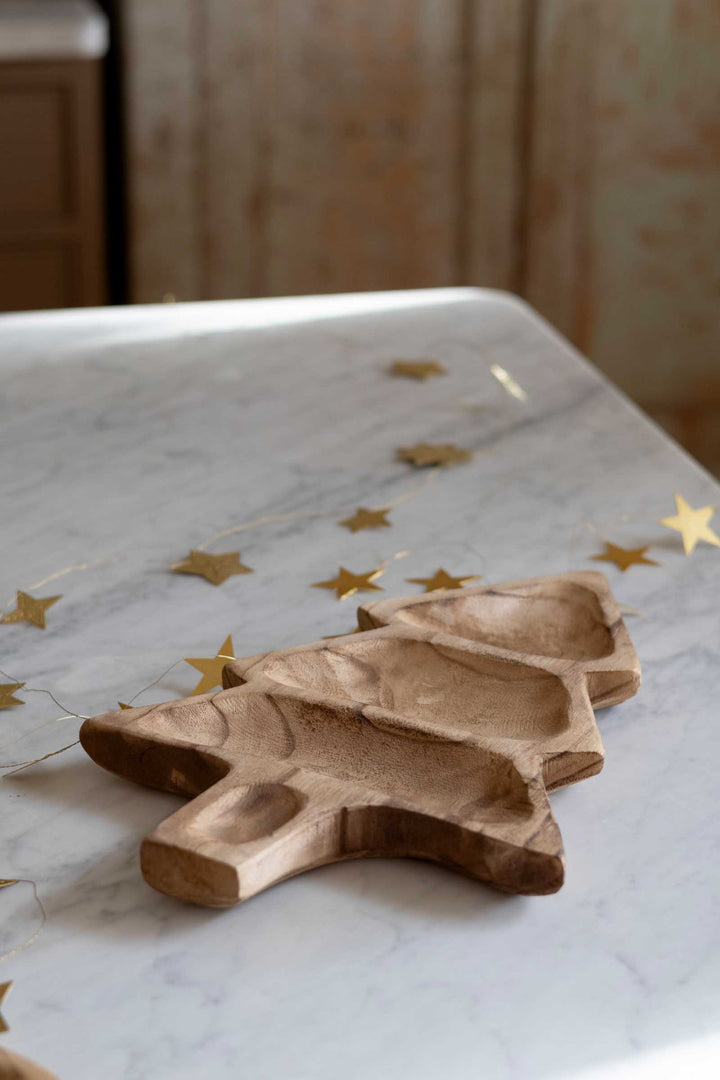 A carved wooden Christmas tree-shaped serving tray with three compartments, placed on a white marble tabletop with scattered gold star decorations.