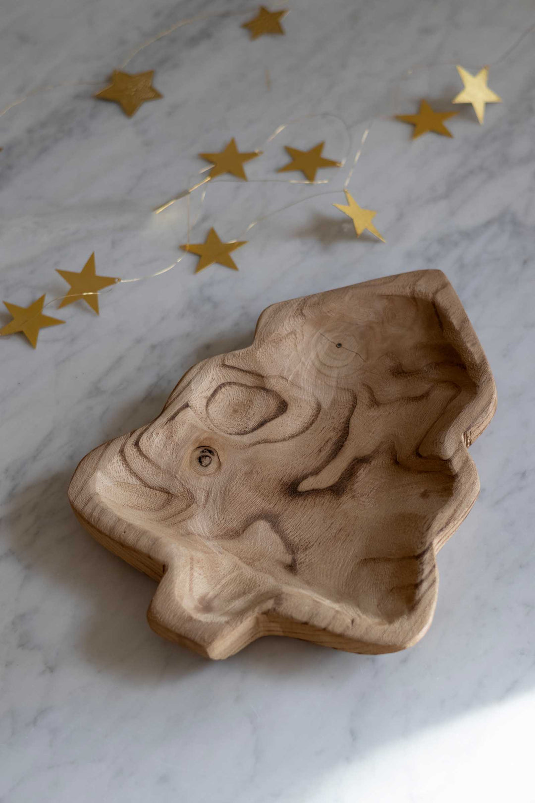 A hand-carved wooden Christmas tree-shaped tray with natural wood grain, placed on a white marble surface with gold star string lights in the background.