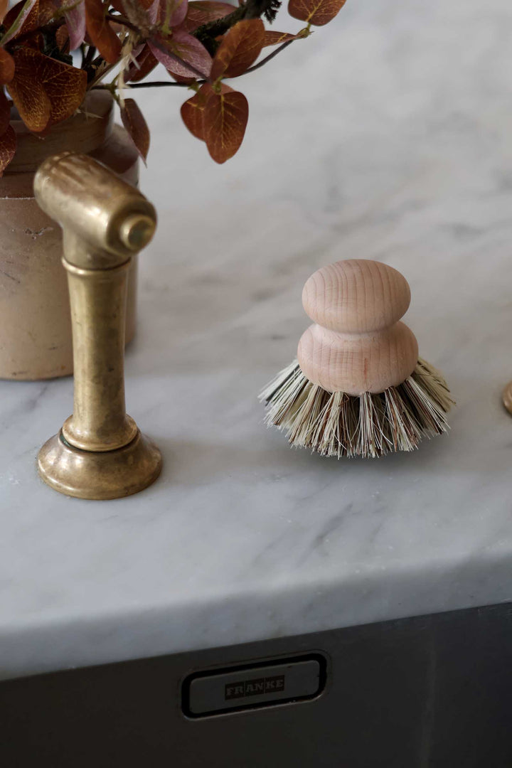 A close-up of a double wooden-handled cleaning brush with natural fiber bristles on a white marble countertop next to a brass faucet.
