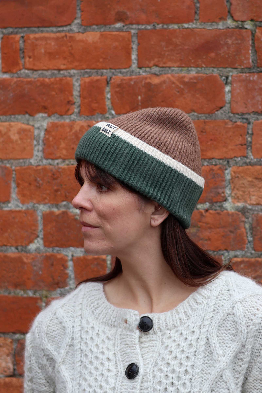 A woman wearing a brown and dark green striped knit beanie with a white stripe, standing in front of a red brick wall.