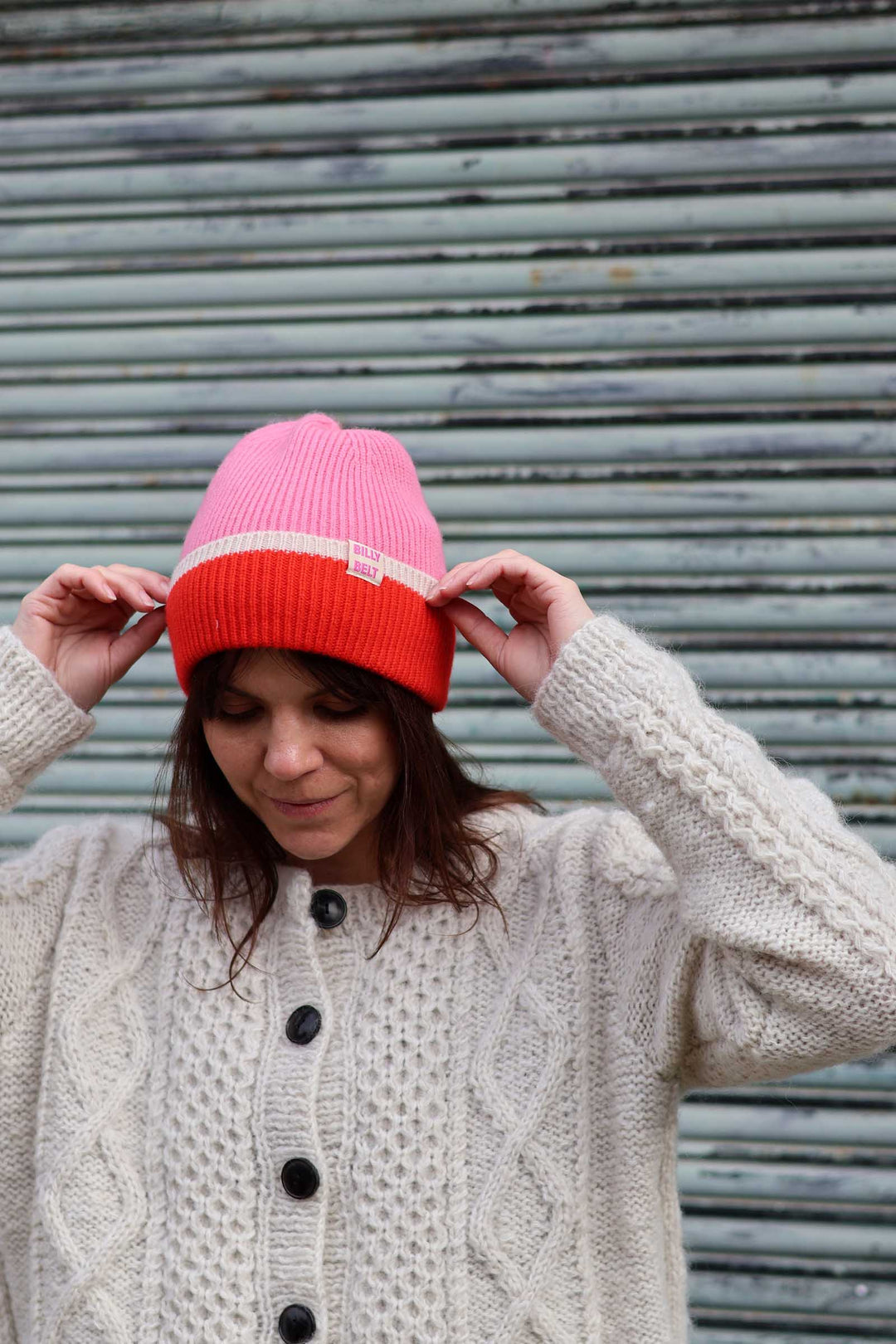 A woman wearing a pink and orange knit beanie with a white stripe, adjusting it with both hands while wearing a cream cable-knit cardigan in front of a gray metal shutter.