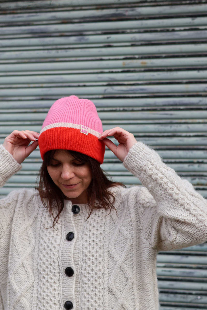 A woman wearing a pink and orange knit beanie with a white stripe, adjusting it with both hands while wearing a cream cable-knit cardigan in front of a gray metal shutter.