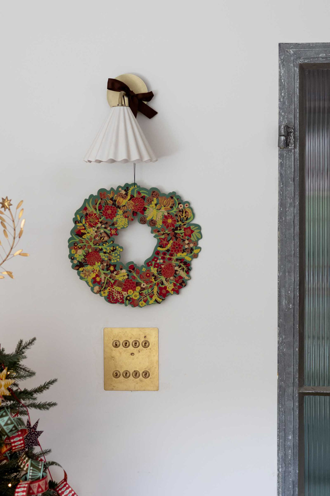 A vintage-style multicolor floral wreath hangs on a white wall below a white pleated wall lamp with a dark brown bow. A gold-toned wall plate is visible below the wreath, with part of a decorated Christmas tree and a gray-framed window on the right.