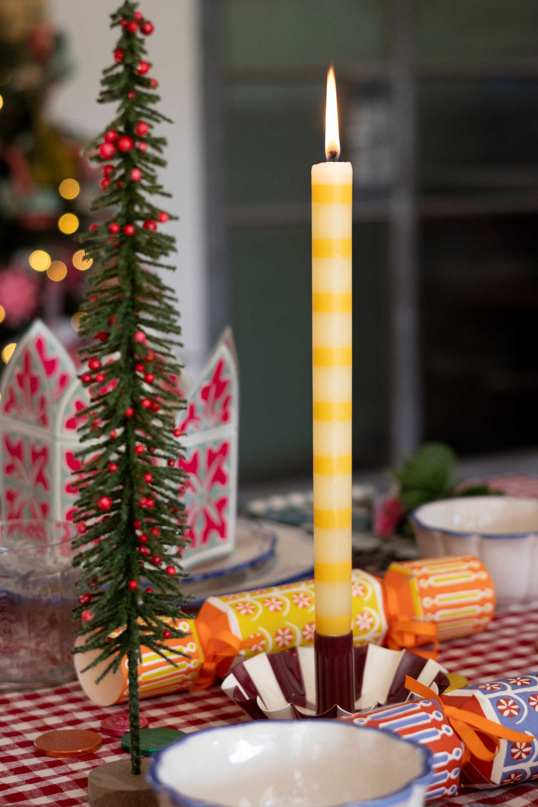 A lit yellow-and-white striped taper candle stands upright in a decorative holder on a table, surrounded by holiday decorations including a small evergreen tree with red ornaments, patterned napkins, and wrapped crackers in a festive indoor setting.
