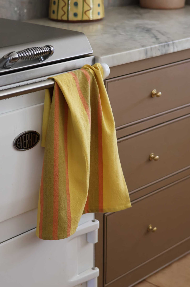 A yellow kitchen towel with orange stripes hanging from the handle of a white Aga stove in a kitchen setting.