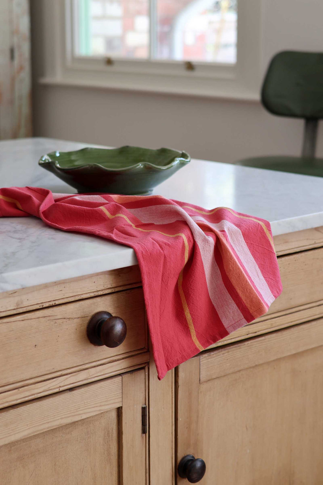 A coral pink striped kitchen towel draped over a light wood kitchen island with a white marble countertop, next to a green ceramic bowl.