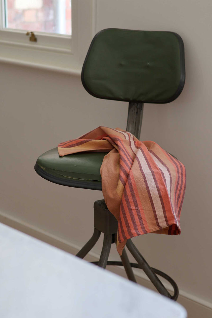 An olive green upholstered stool with a matching backrest and gray metal legs, shown indoors against a beige wall near a window. A peach and orange striped fabric is draped over the seat.