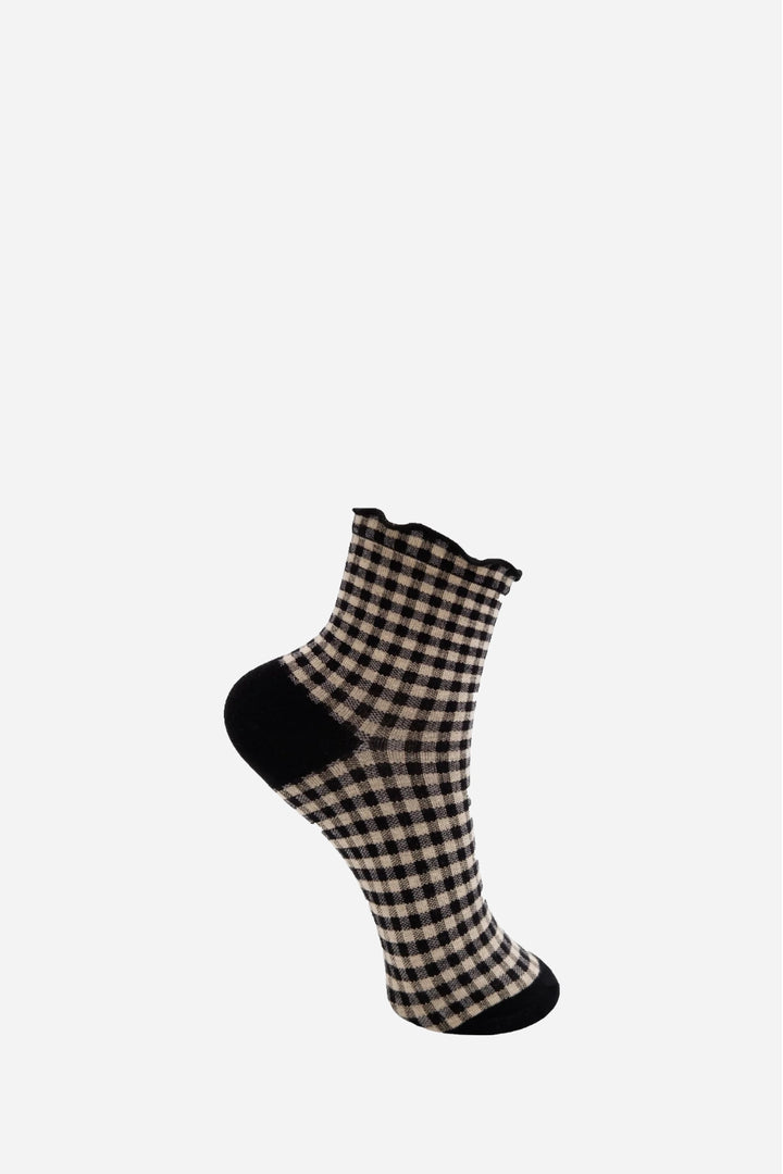 A single ankle sock with a beige and black gingham check pattern, featuring solid black heels, toes, and a ruffled cuff, shown against a plain light gray background.
