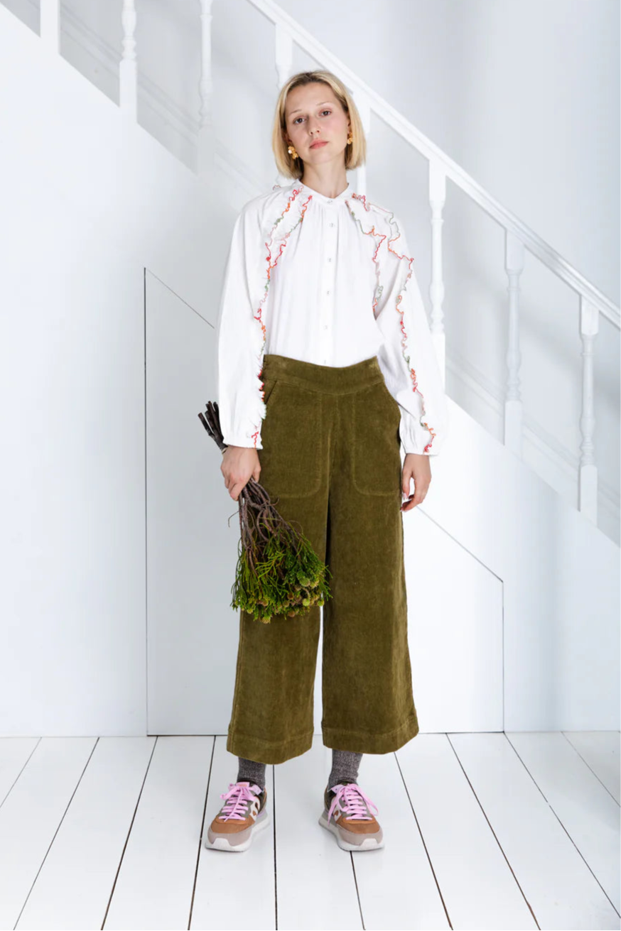 Person wearing a white blouse with red patterns and green pants in a minimalistic room.