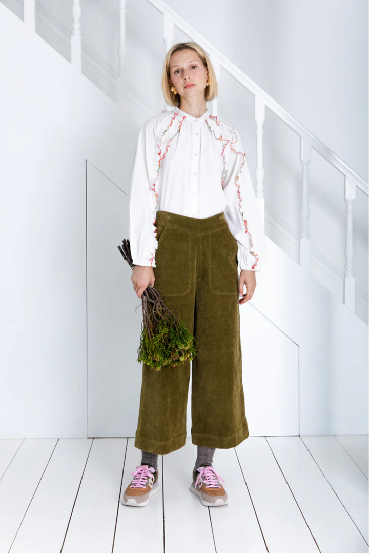 Person wearing a white blouse with red patterns and green pants in a minimalistic room.