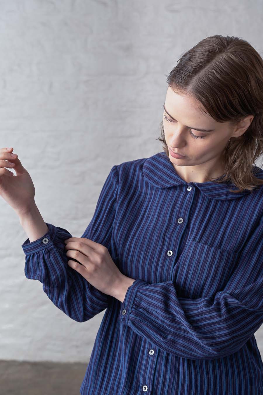 A woman wearing a dark blue and navy vertically striped long-sleeve button-up shirt, adjusting the cuff of her sleeve against a light gray textured wall.