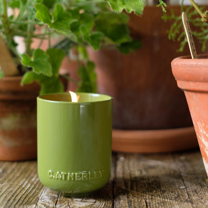 Green candle with 'C. Atherley' branding on a wooden surface with potted plants in the background.